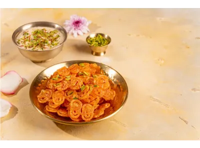 Golden plate of coin jalebi with a bowl of rabdi, sliced pistachios, and flowers on a beige background.