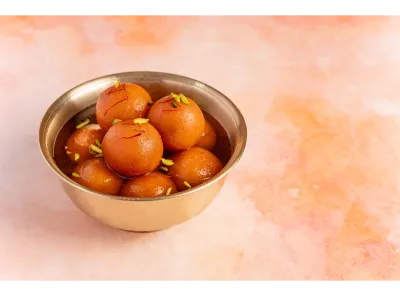 Bowl of delicious Indian sweet gulab jamun garnished with pistachio and saffron on a pink background.