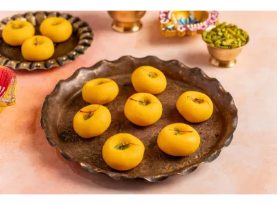 Kesar Pedas garnished with saffron and pistachios on a bronze plate with festive decor in the background.