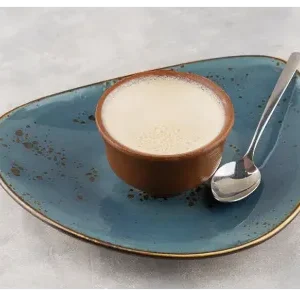 Traditional Mishti Doi served in an earthen pot on a blue ceramic plate with a spoon beside it on a grey textured surface.