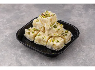 Pieces of plain Kalakand garnished with chopped pistachios, stacked in paper cups on a black tray