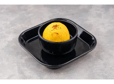 Single yellow Raj Bhog sweet placed in a black bowl and tray, topped with saffron strands