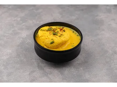 Reshmi Rasmalai served in a black bowl, soaked in saffron milk and garnished with herbs on a textured grey surface.