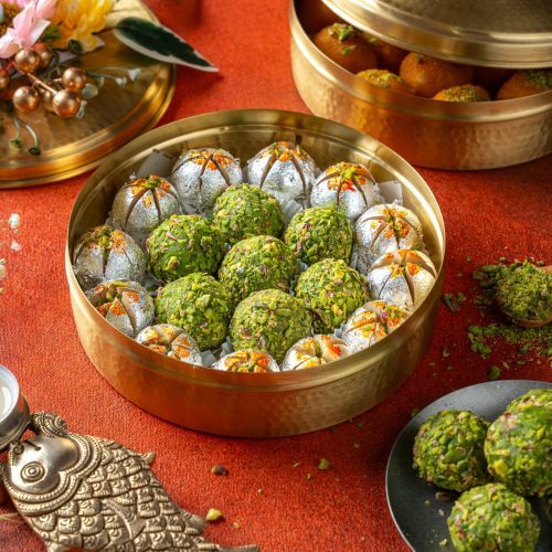 Brass round box filled with pistachio and silver-coated Indian laddus, placed on a festive red cloth with traditional decor.