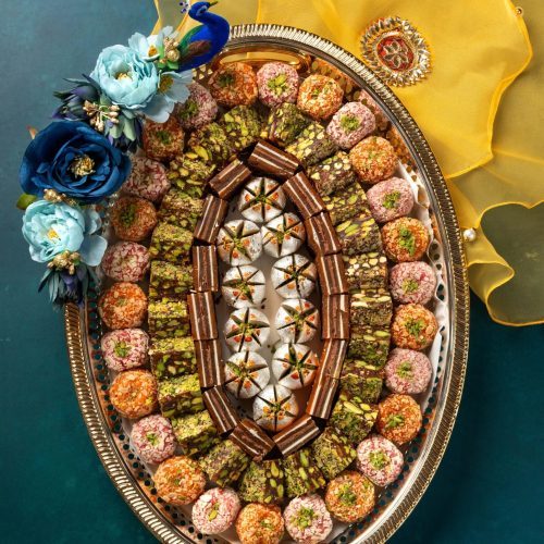 Decorative silver tray with an assortment of colorful Indian sweets, adorned with flowers and festive yellow cloth on the side