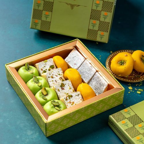 Green gift box with assorted mawa sweets including green apple-shaped peda, barfi, and yellow sweets